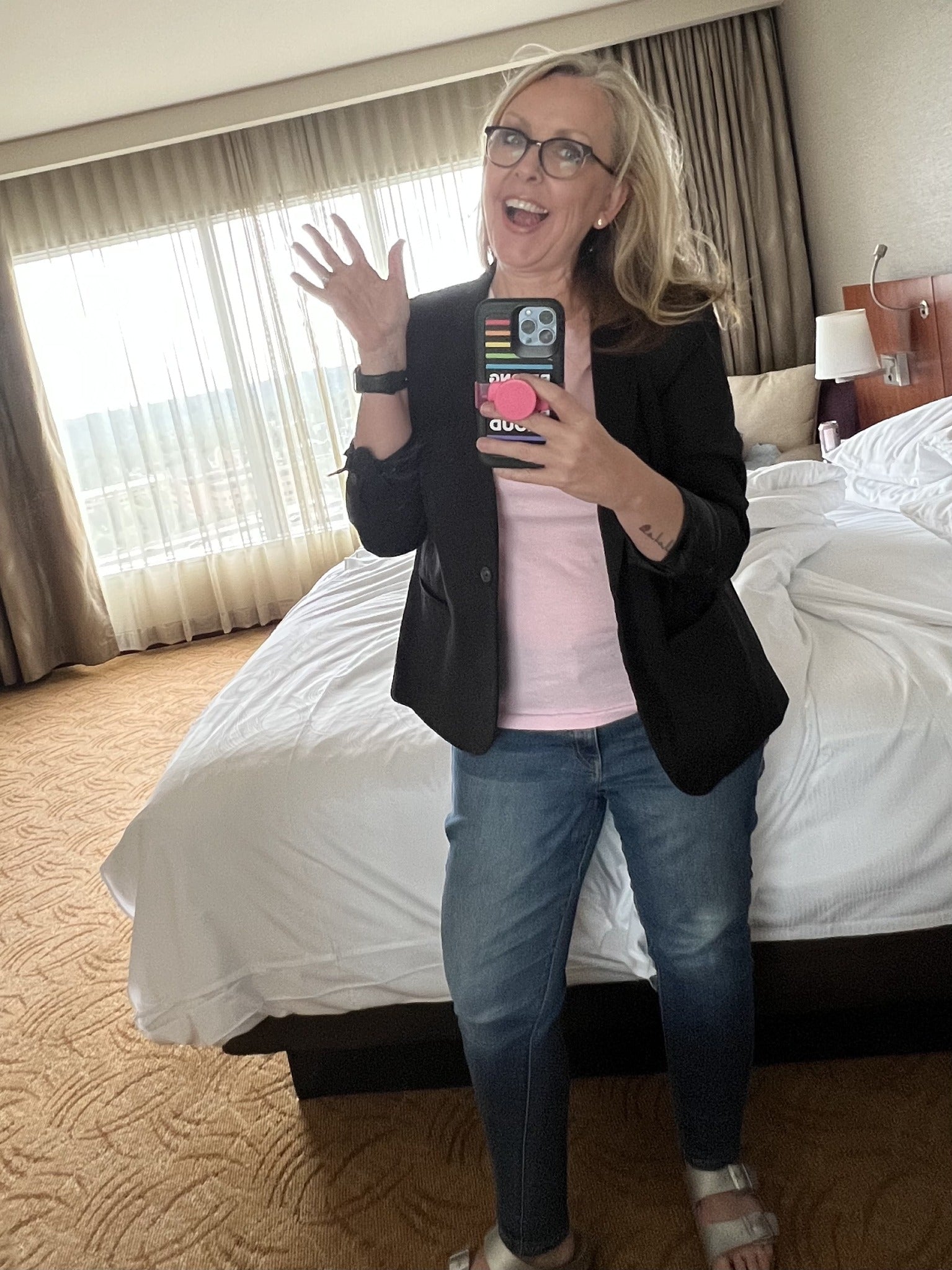 Brenda smiling and waving in a mirror selfie, wearing a black blazer, pink top, and jeans
